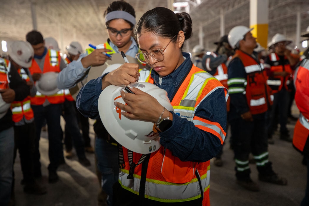 Trabajadora con chaleco naranja firma un casco blanco durante la Semana de la Seguridad.