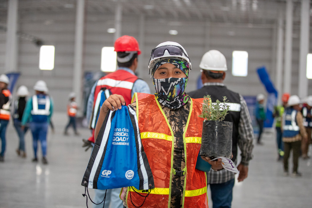 Trabajador con chaleco naranja muestra una mochila azul y una planta en maceta.