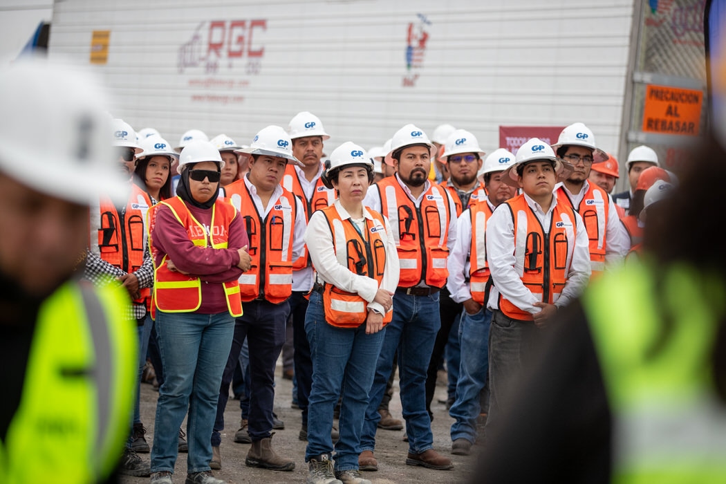Fila de trabajadores con chaleco naranja y casco blanco escucha en la obra.