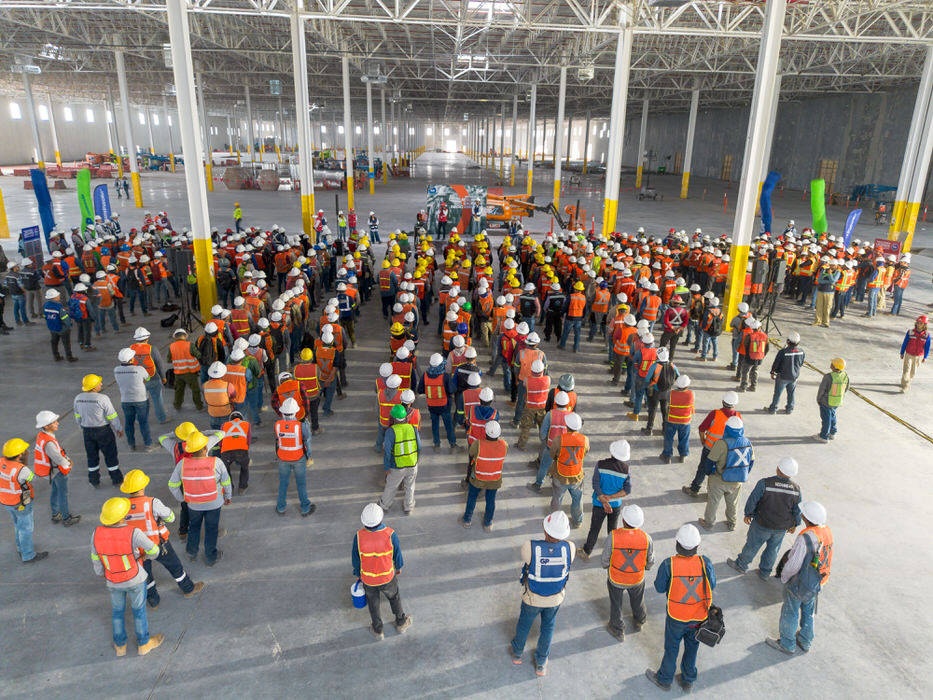 Gran grupo con cascos y chalecos naranja se reúne bajo la estructura de una nave.