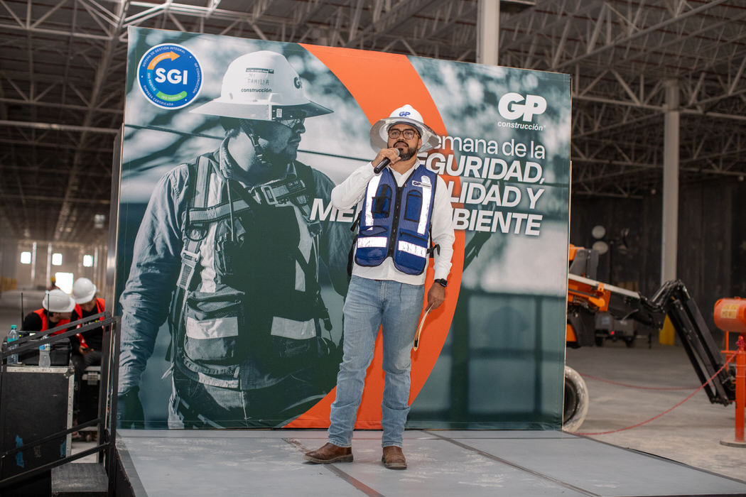 Ponente con chaleco azul habla por micrófono frente al letrero de la Semana de la Seguridad.