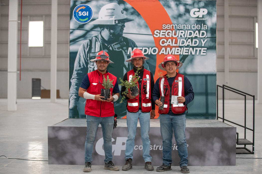 Tres trabajadores con chaleco rojo posan con plantas frente al letrero de la Semana de la Seguridad.