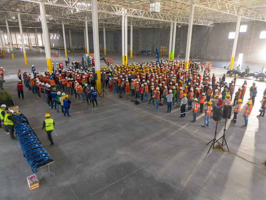 Asamblea en nave industrial, con bocinas, chalecos apilados y columnas de base amarilla.