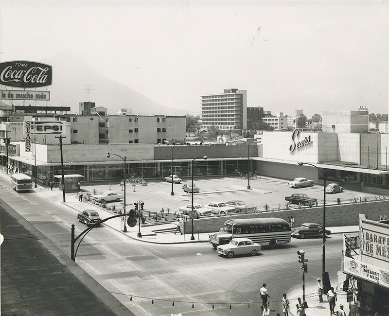 Tienda Sears con estacionamiento, autos de época y un anuncio de Coca-Cola.