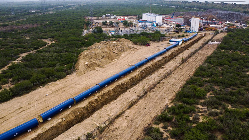 Tubería azul del Acueducto El Cuchillo sobre una zanja, con tanques al fondo.