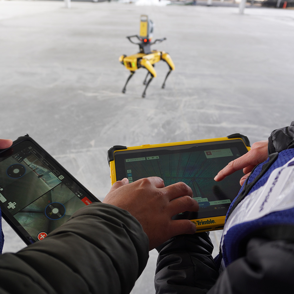 Manos operan tabletas Trimble mientras un robot cuadrúpedo amarillo recorre la losa.