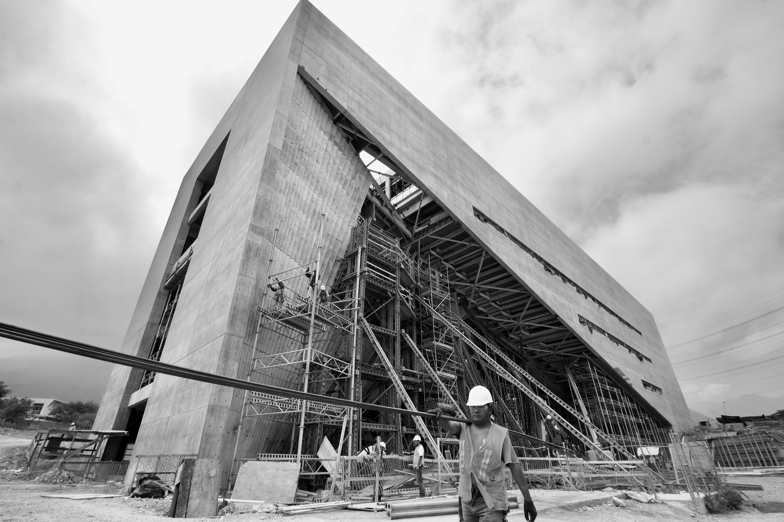 Trabajador con casco frente a un edificio de concreto en obra, con andamios.