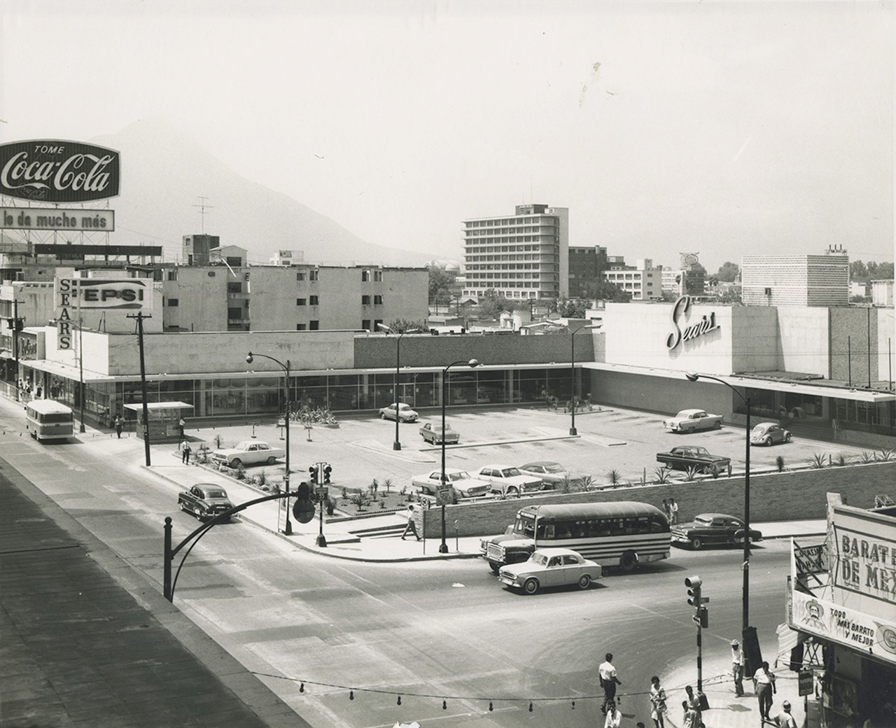 Centro comercial con estacionamiento, anuncios y el cerro al fondo, en toma histórica.