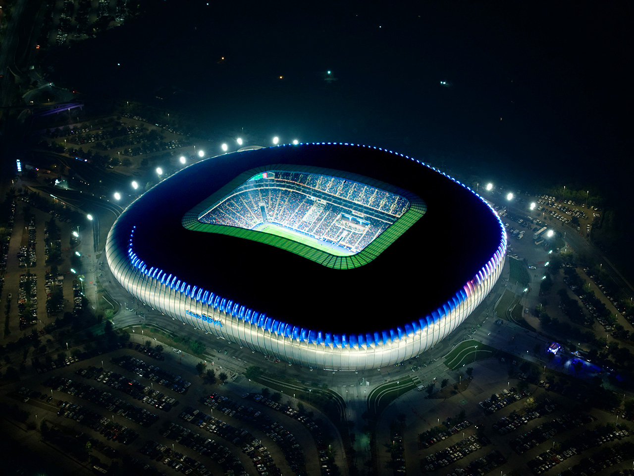 Estadio iluminado de noche, visto desde el aire, rodeado de estacionamiento.