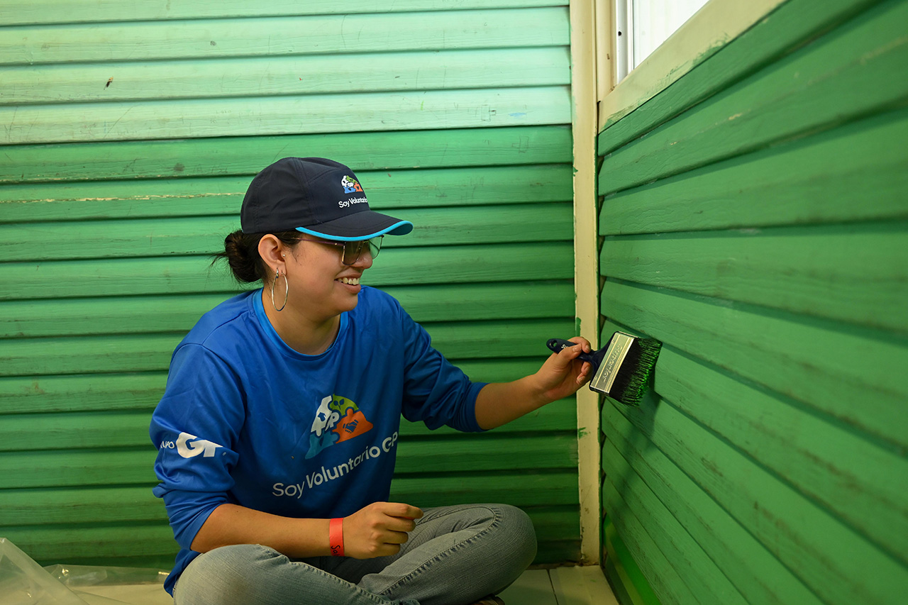 A volunteer from Grupo GP is painting a sheet metal wall green while sitting on the floor.