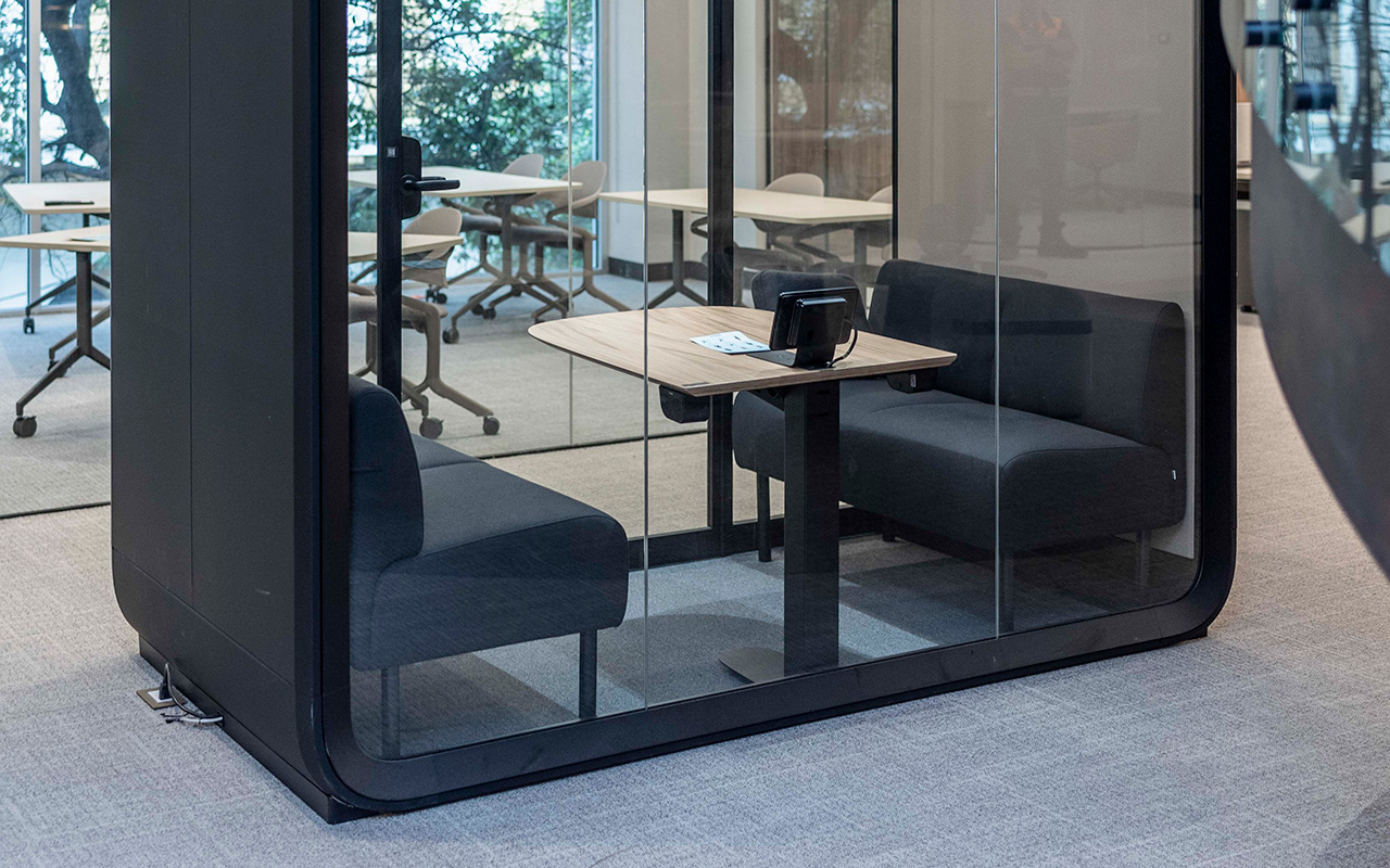 Cabina acristalada con dos sofás y una mesa, frente a las estaciones de trabajo.