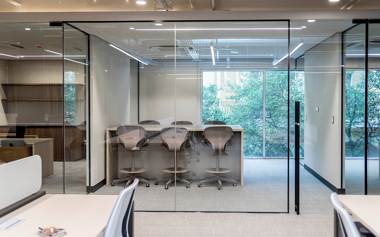 Sala acristalada con cuatro bancos altos frente a ventanales con árboles.