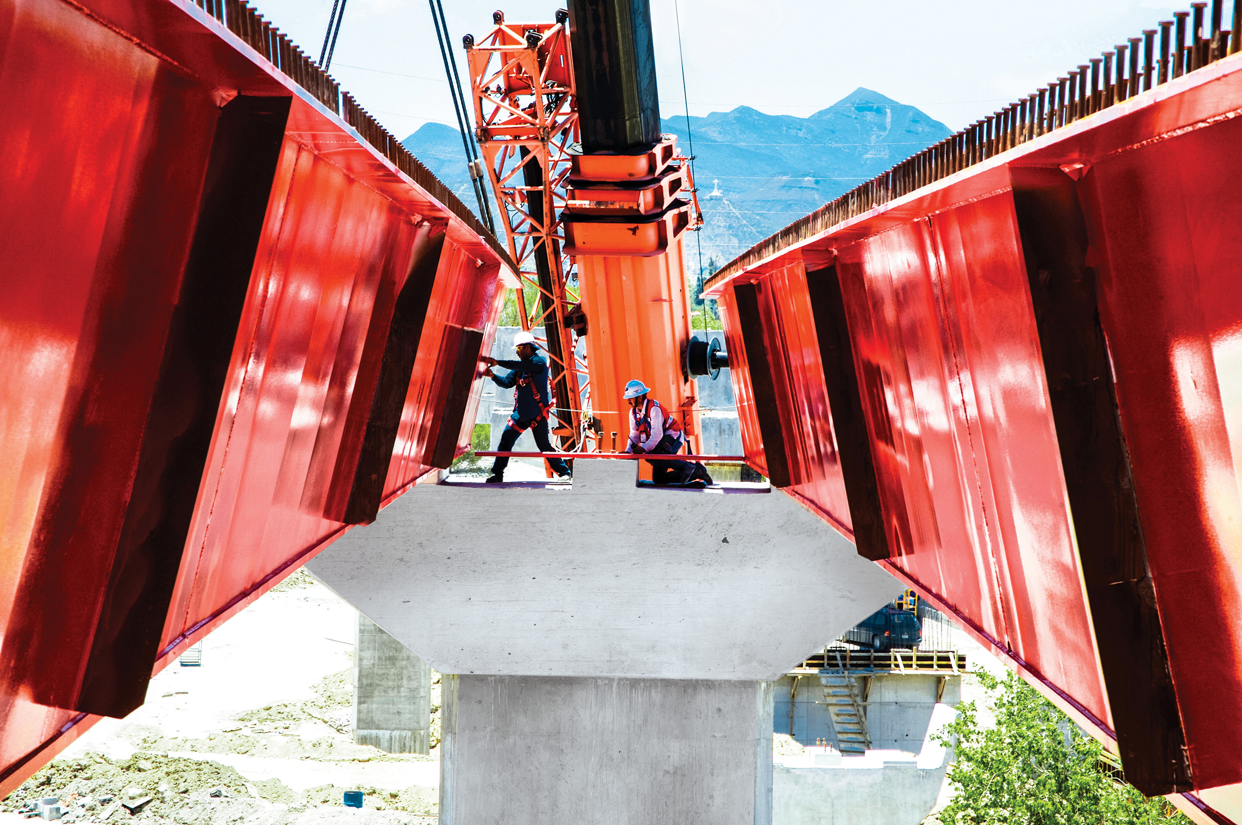 Trabajadores montan vigas rojas de un puente, con la sierra al fondo.