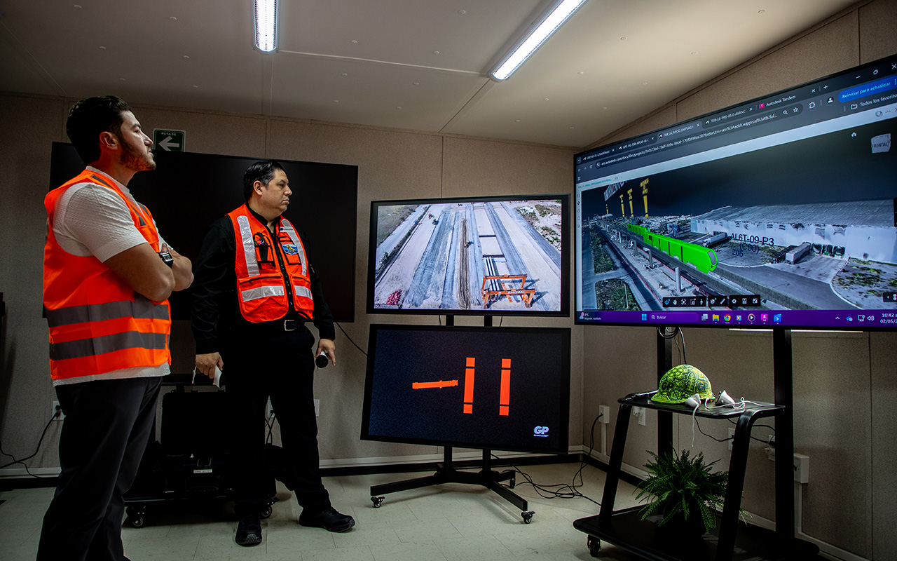 Dos supervisores observan un recorrido aéreo y un plano en pantallas de la sala.