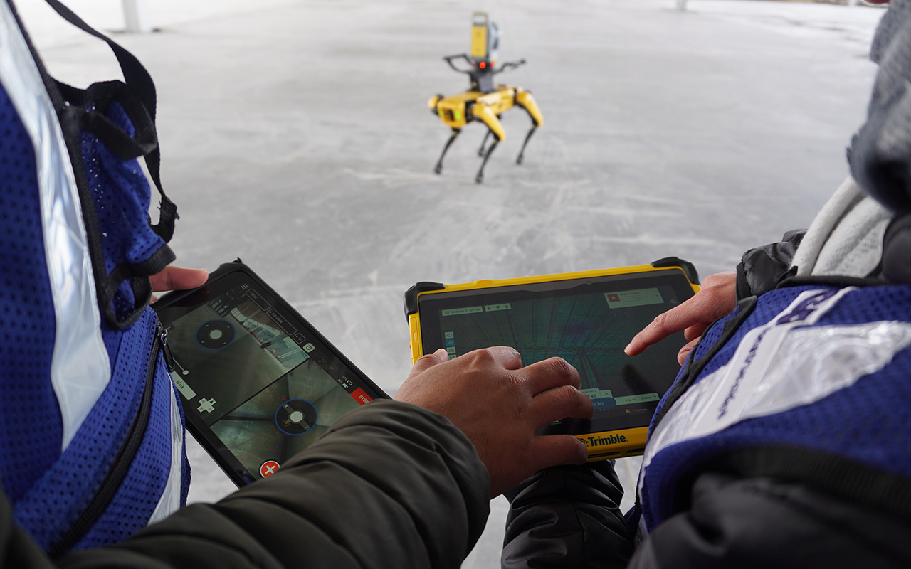 Dos personas con chaleco azul guían por tableta a un robot cuadrúpedo en la losa.