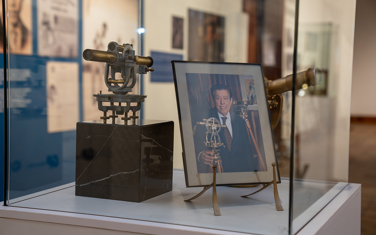 Retrato de un hombre con traje y corbata sosteniendo un instrumento de medición óptica, junto a otro instrumento similar de latón expuestos en una vitrina de museo.