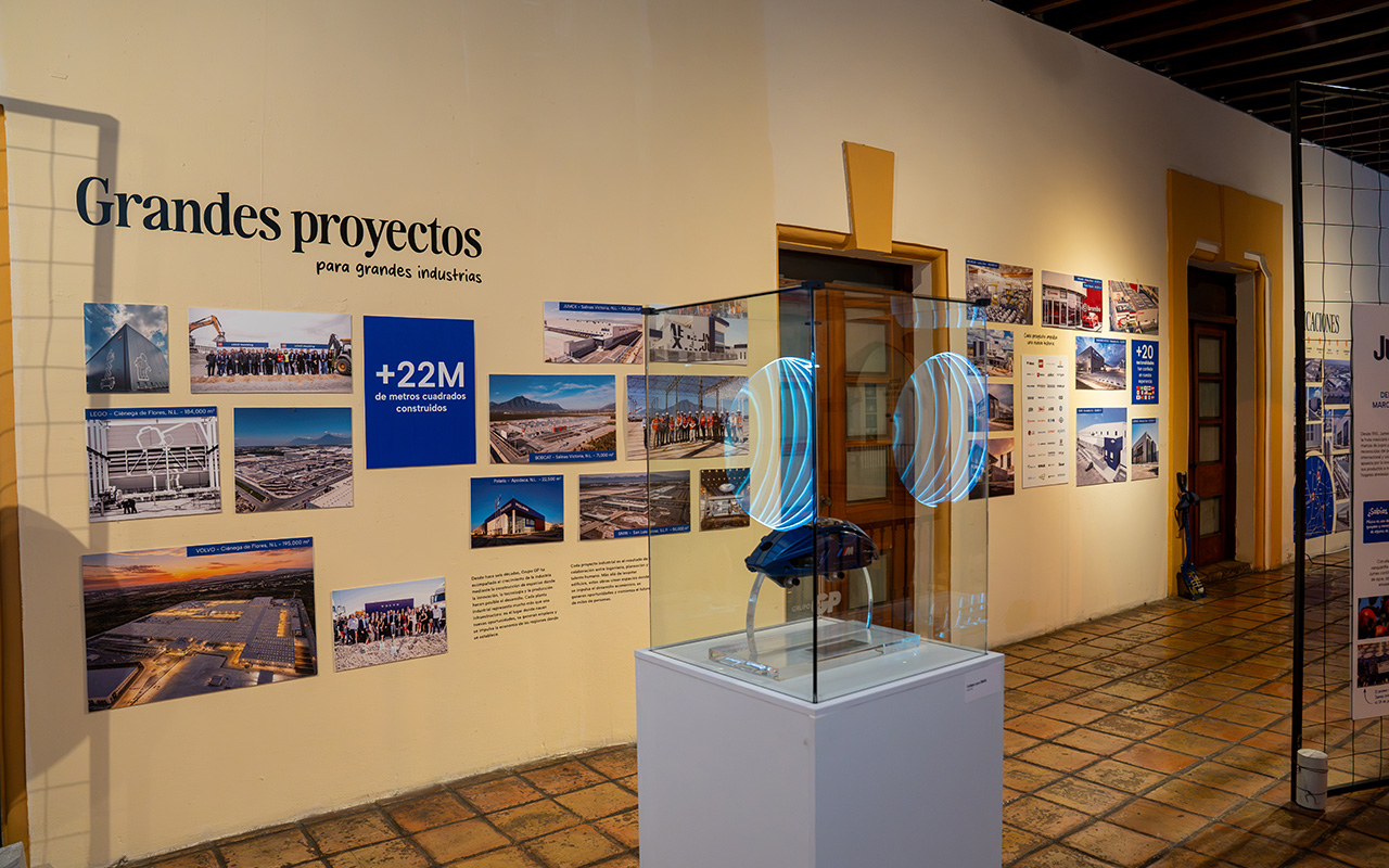 Exhibición en museo con paredes exhibiendo fotos y textos sobre grandes proyectos industriales que suman más de 22 millones de metros cuadrados construidos, con un objeto de arte iluminado dentro de una vitrina de vidrio en el centro.