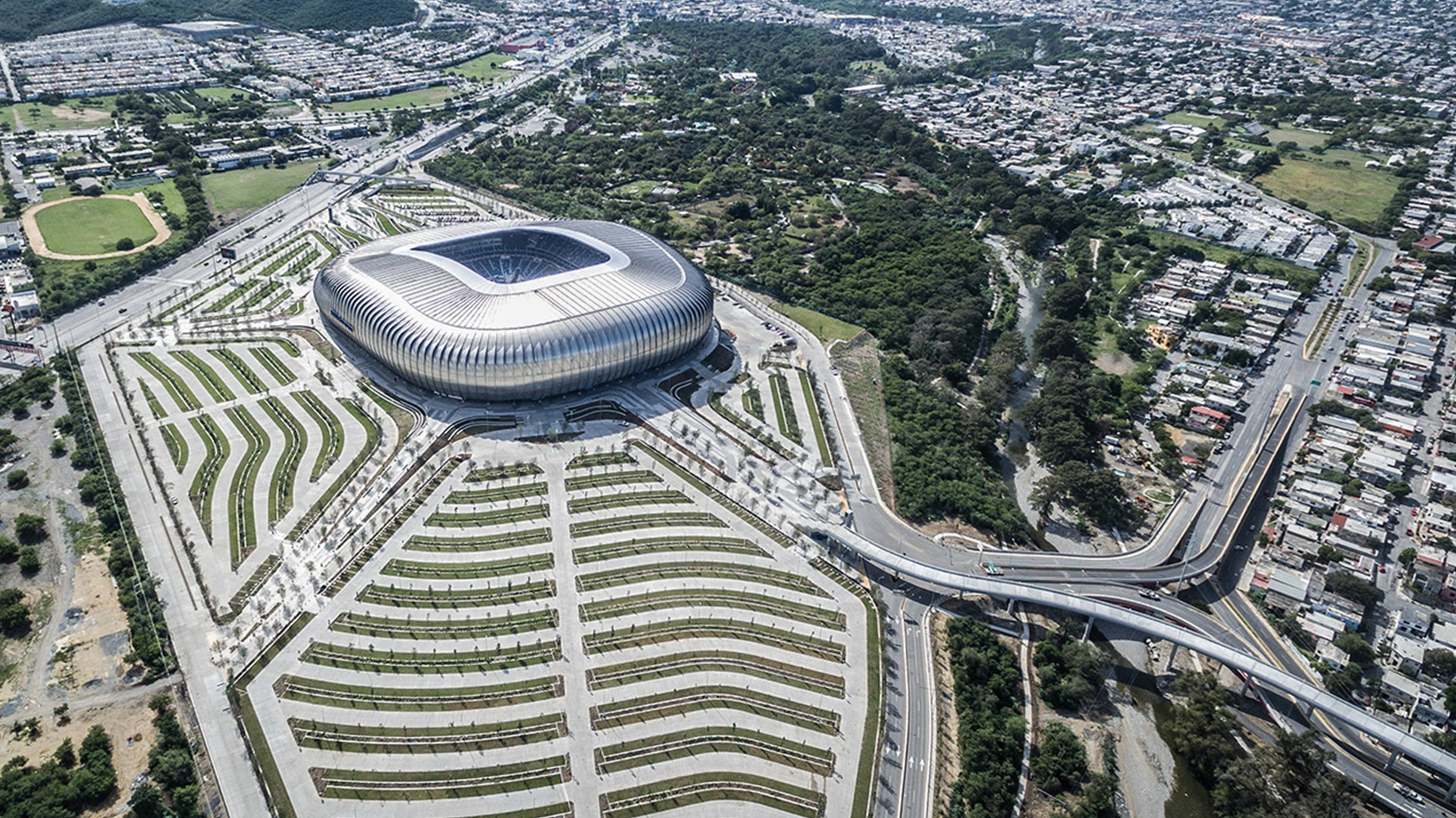 Vista aérea del Estadio BBVA, con jardines ondulados, vialidades y la ciudad alrededor.