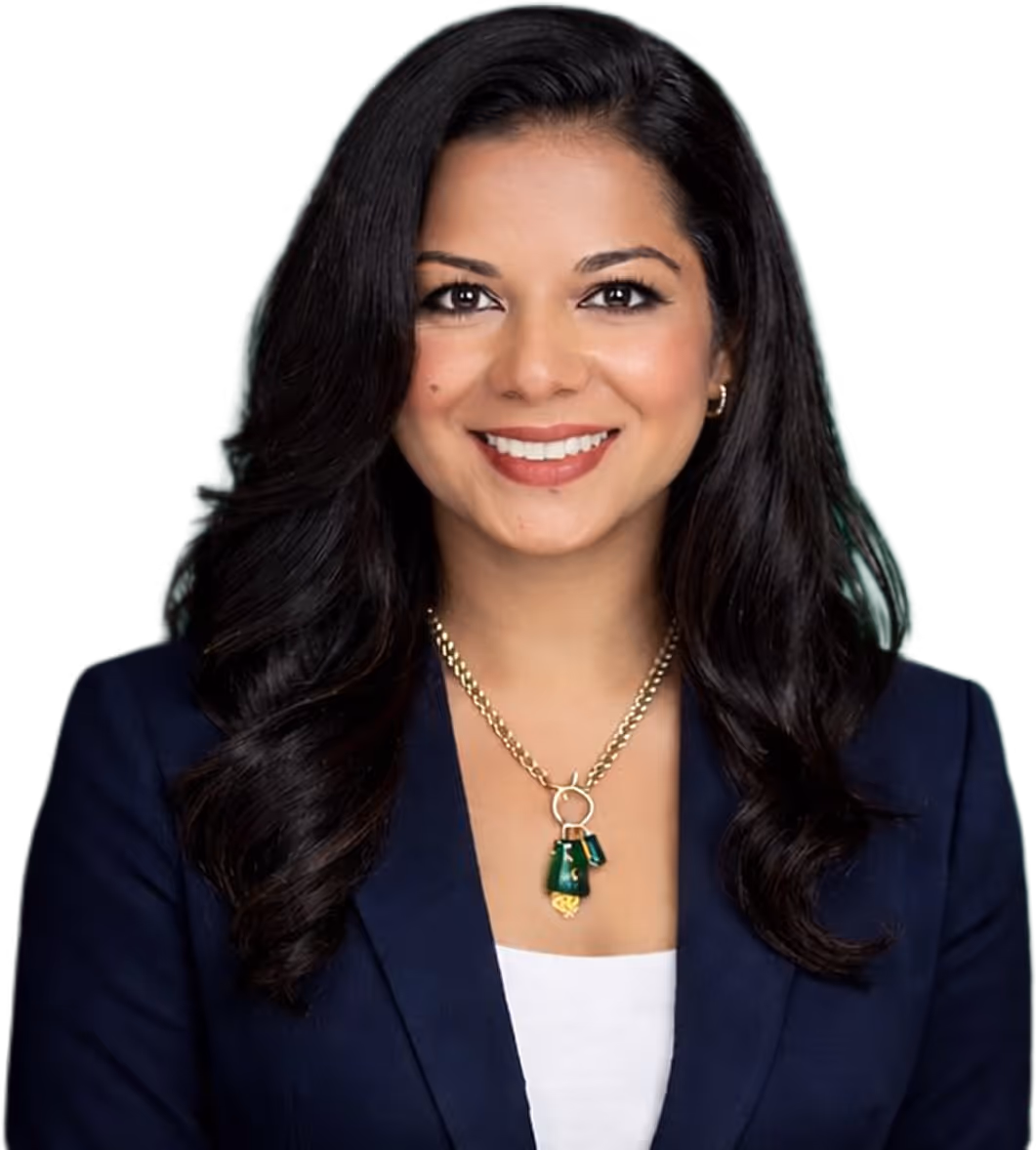 Smiling woman with long dark hair, wearing a navy blazer, white top, and gold necklace with green pendants.