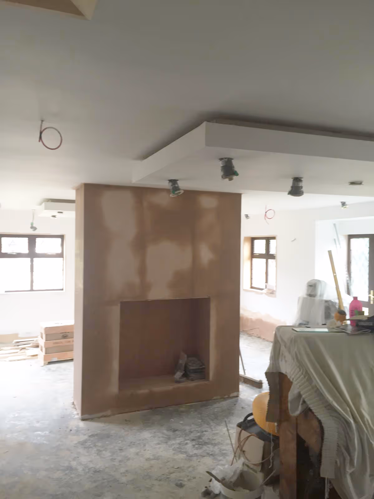 Interior of a room under renovation with an unfinished brown plaster fireplace in the center and construction materials scattered around.