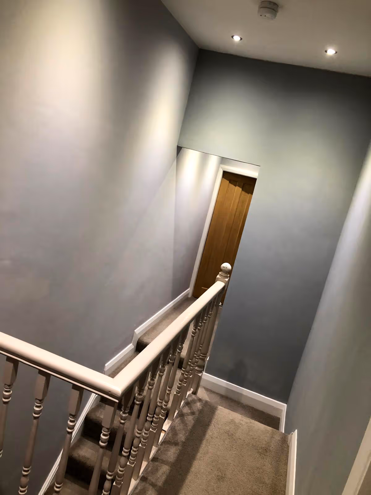 Indoor staircase with beige carpet, white wooden railing, gray walls, and a closed wooden door at the hallway end.