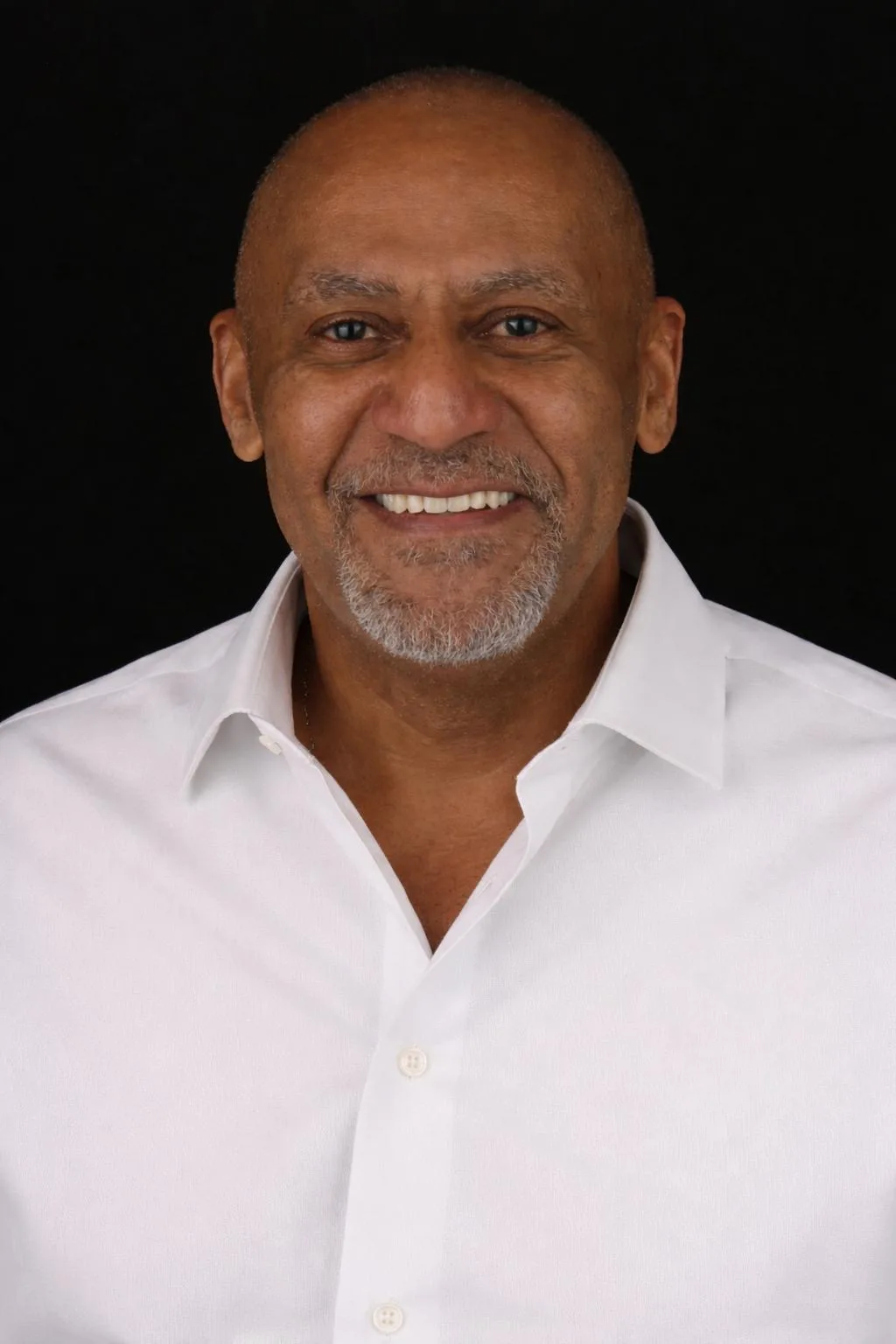 Smiling middle-aged man with a shaved head and gray goatee wearing a white button-up shirt against a black background.