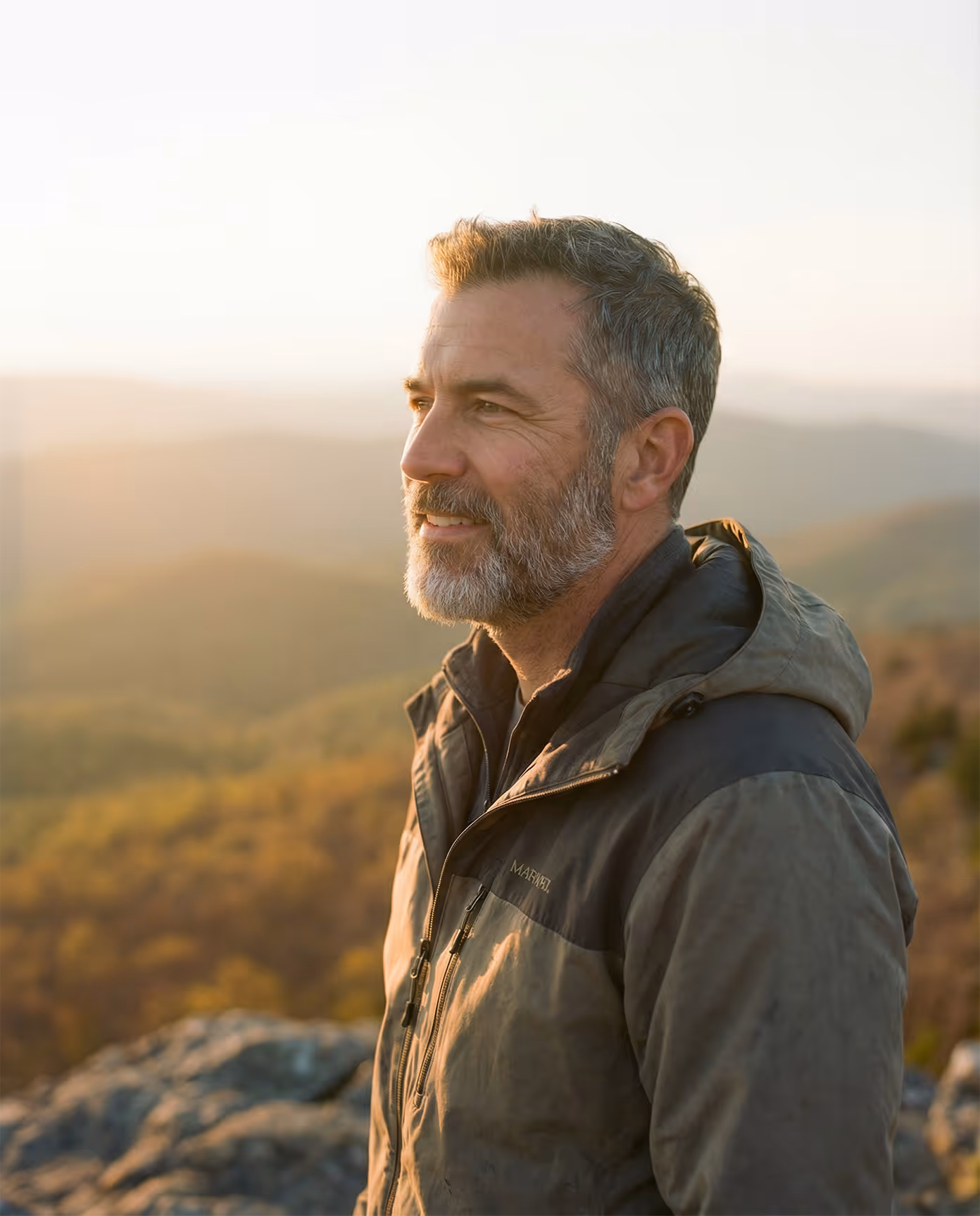 Smiling middle-aged man with gray beard wearing jacket outdoors at sunset with mountains in background.