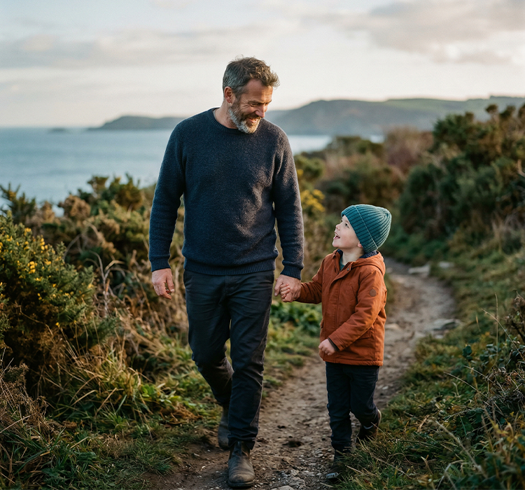 Man and child holding hands walking on a coastal path with greenery and water in the background.