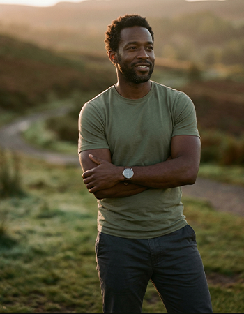Man in green shirt and watch standing outside on a grassy trail at sunset, looking to the side.