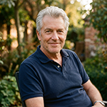 Older man with gray hair wearing navy blue polo sitting outdoors with greenery in background.