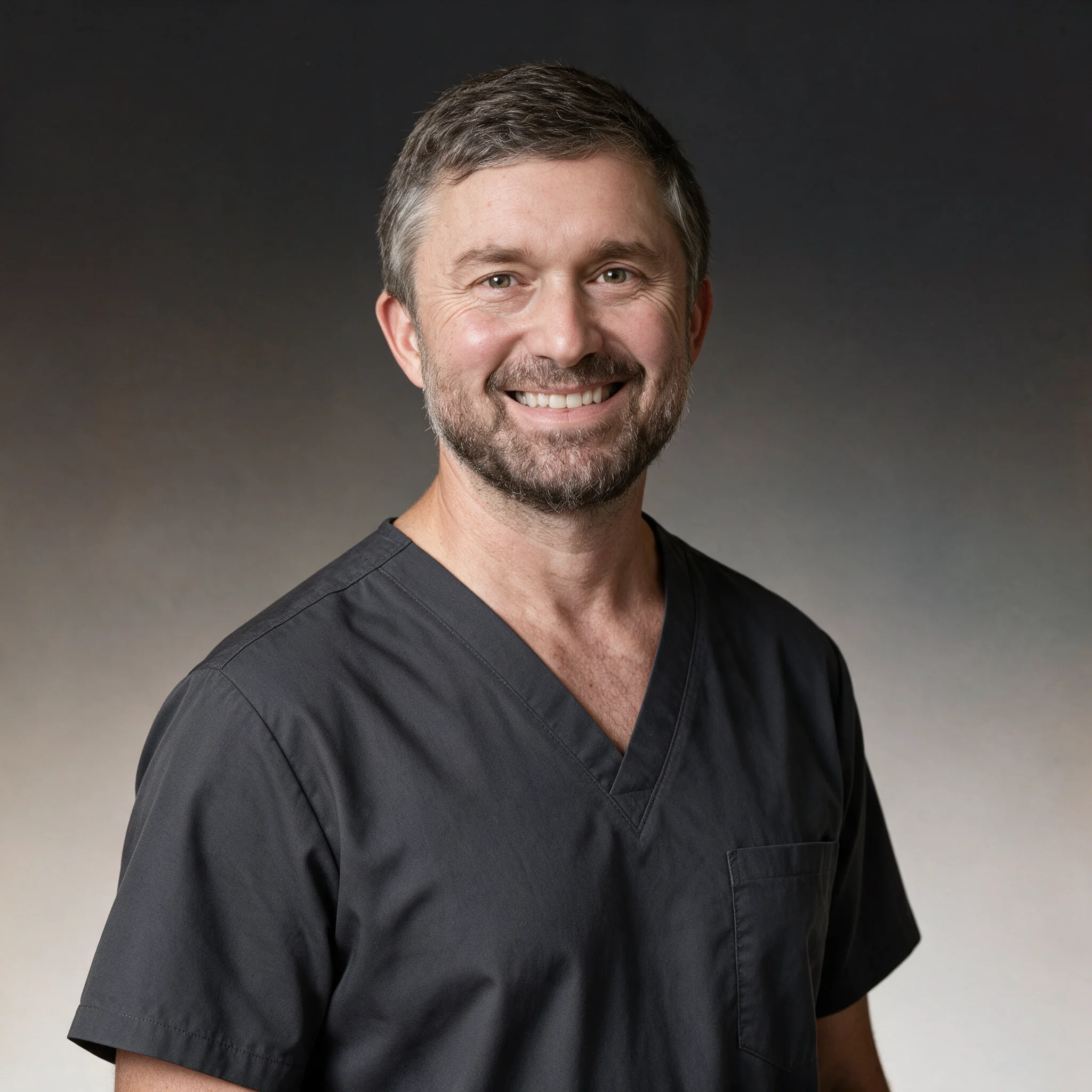Smiling middle-aged man with short graying hair and beard wearing dark gray medical scrubs against a gradient gray background.