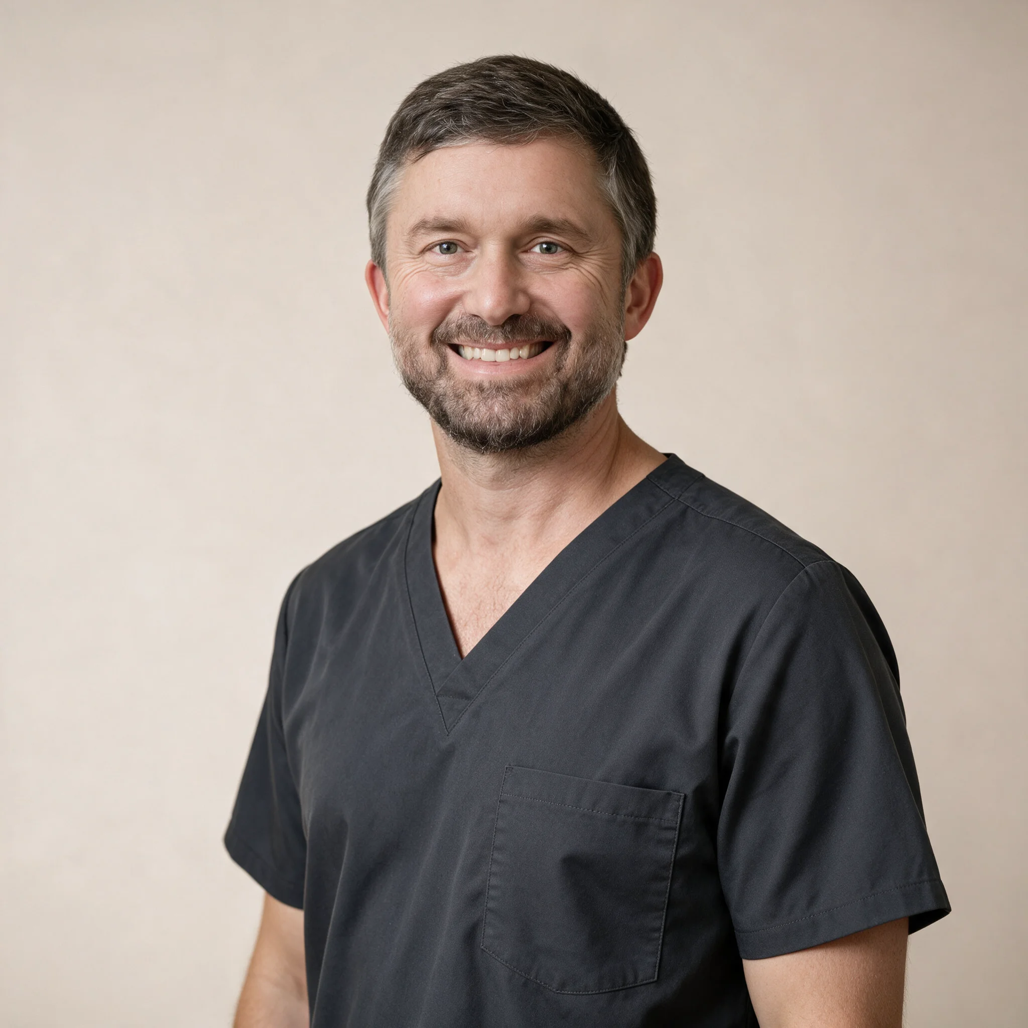 Smiling man with short brown hair and beard wearing a dark gray medical scrub top against a neutral light background.