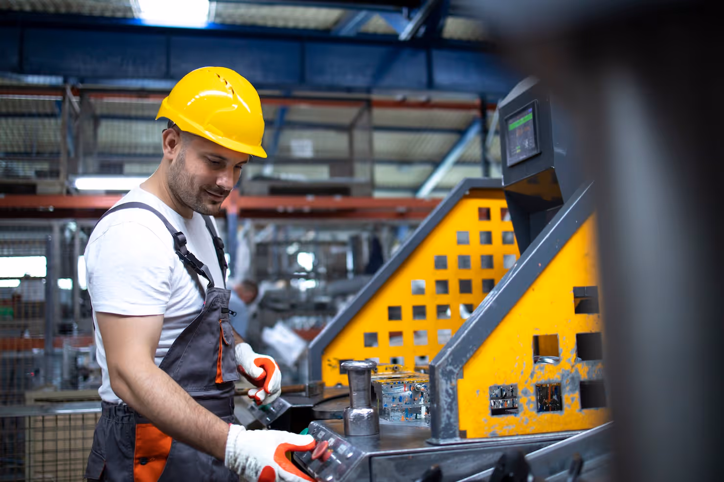 Worker wearing yellow hard hat and gloves operating industrial machinery with metal parts in a factory.
