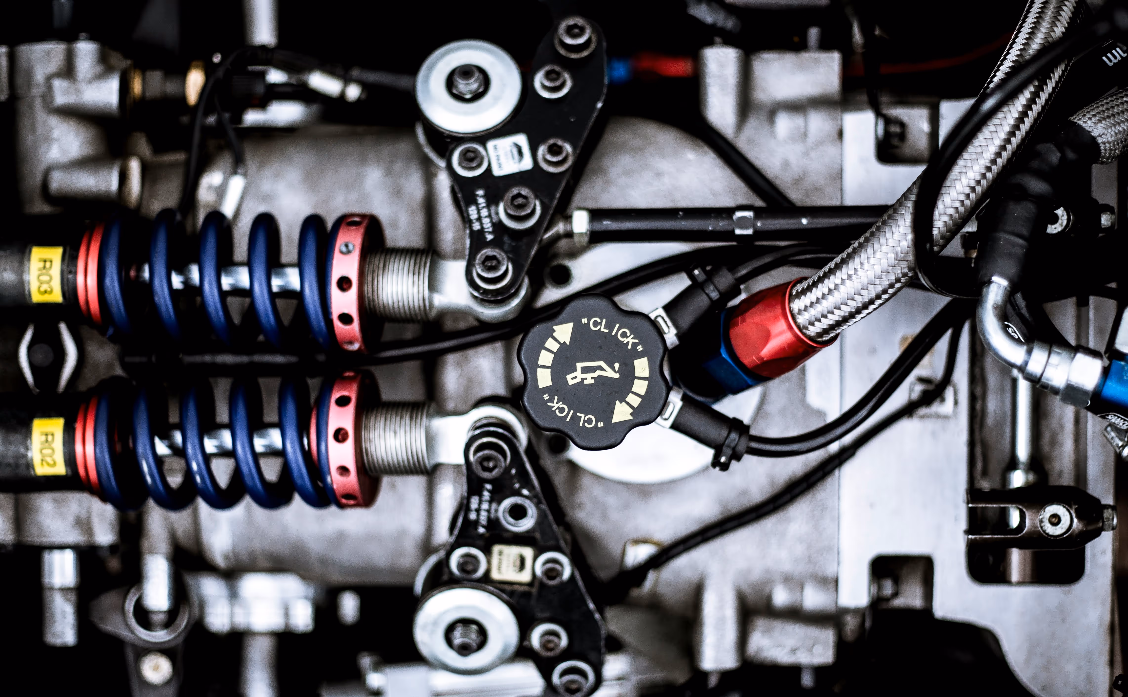 Close-up of a mechanical engine component with coiled springs, bolts, hoses, and a central black cap labeled 'CLICK'.