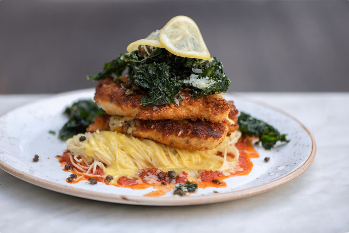 Plate with breaded and fried chicken cutlets on creamy pasta, topped with crispy kale and lemon slices.