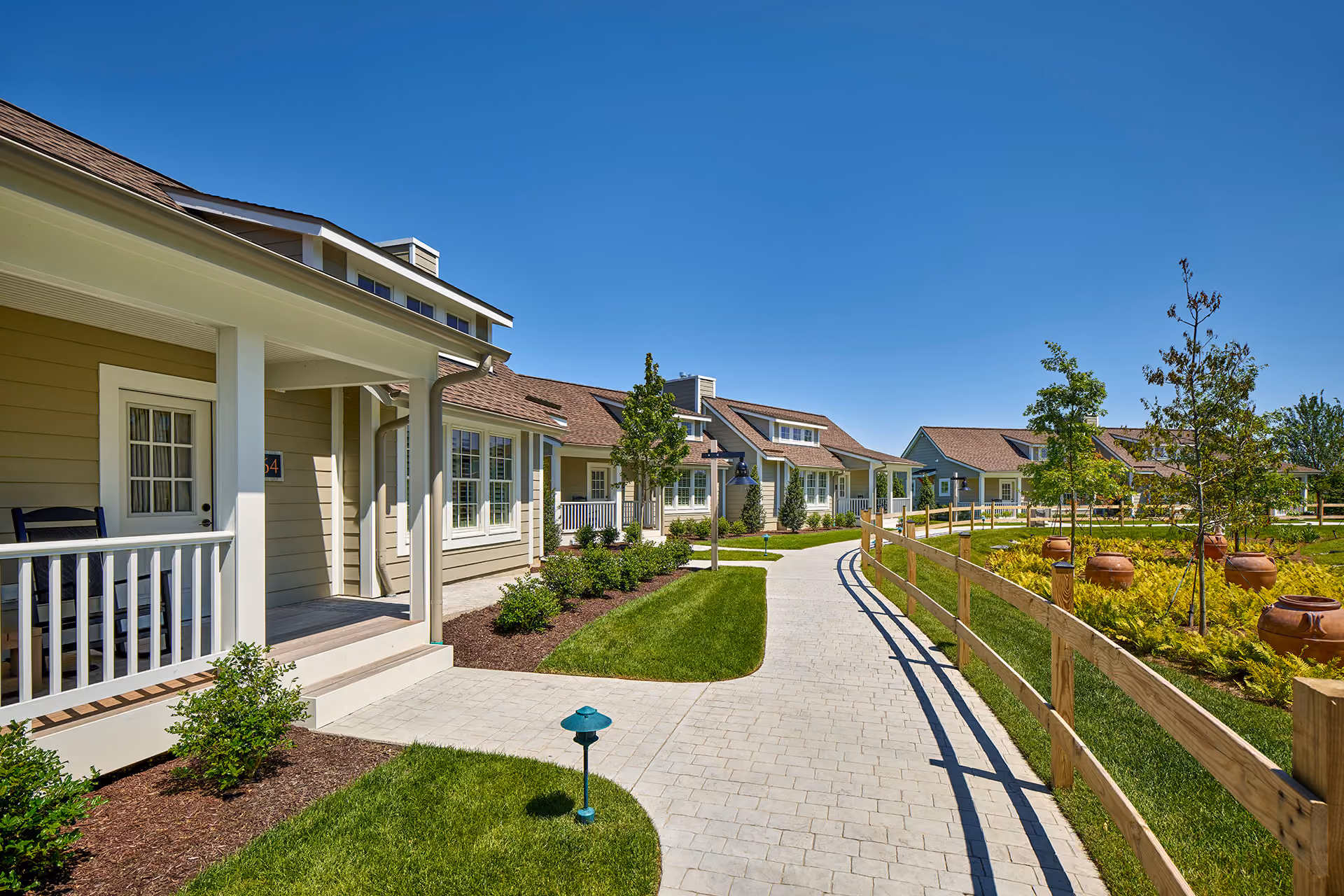 Curved paved walkway alongside beige cottages with front porches, green lawns, and small trees under a clear blue sky.