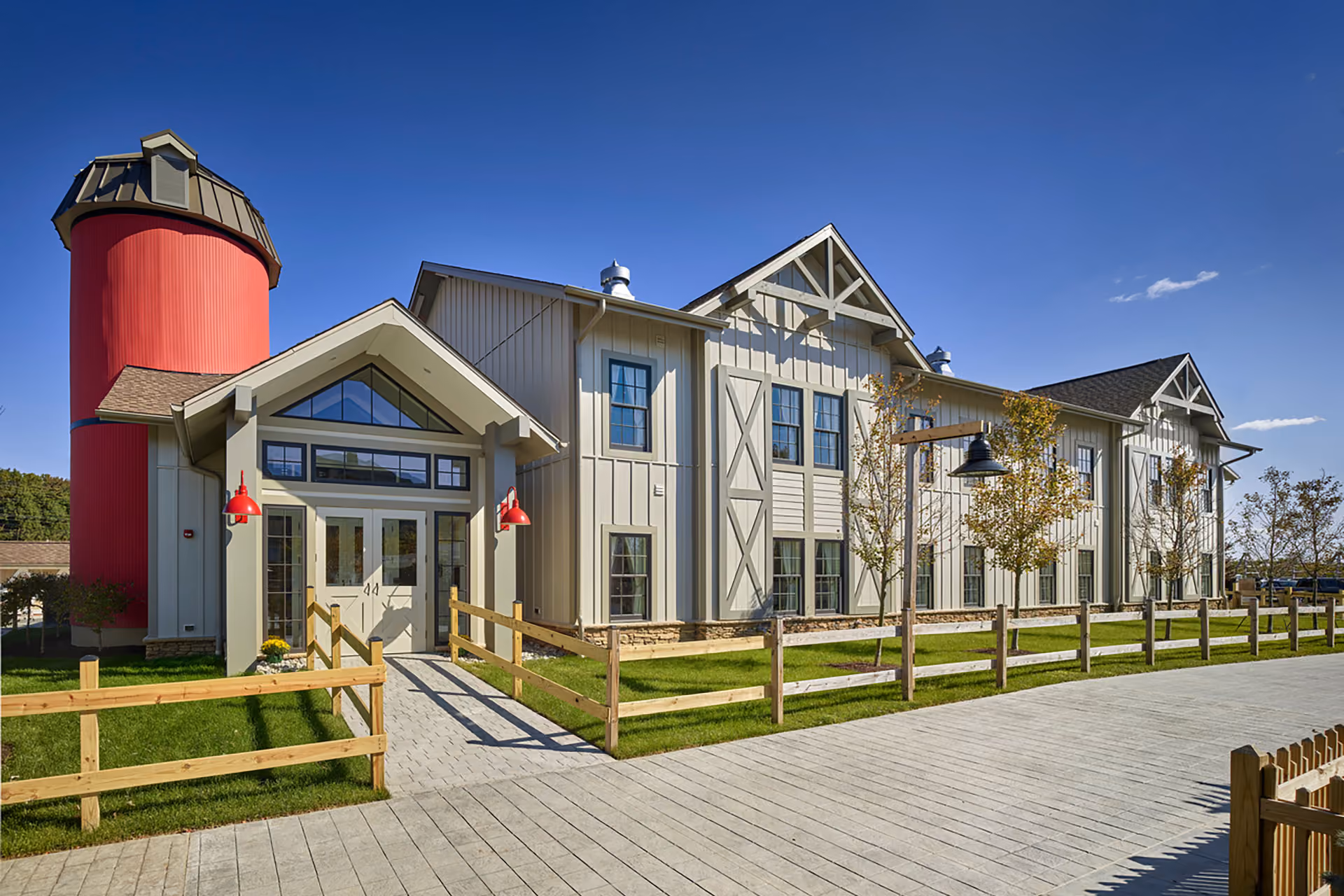 Modern large barn-style building with beige siding, multiple windows, a red silo, red outdoor lamps, and wooden fencing surrounding a paved walkway.