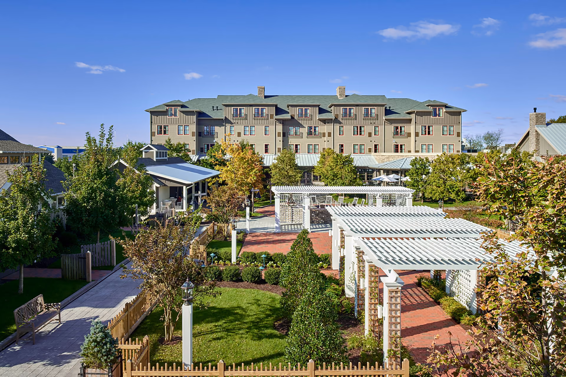 Spacious inn exterior with a large multi-story building, lush gardens, white pergolas, brick pathways, and clear blue sky.