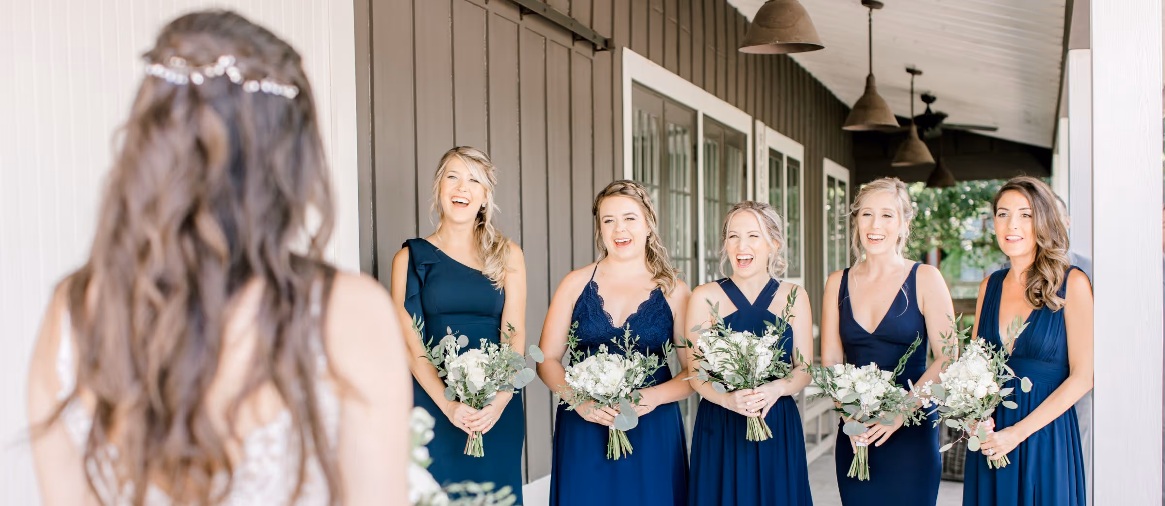 Bride with long wavy hair seen from behind smiling at five bridesmaids in navy blue dresses holding white flower bouquets on a porch.