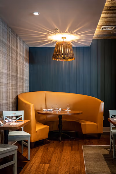 Cozy restaurant corner with a curved mustard-yellow booth, round wooden table, and a hanging wooden light fixture casting a starburst pattern on the ceiling.
