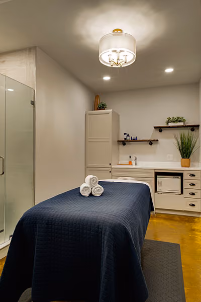 Massage table covered with a dark blue quilted blanket and rolled white towels in a clean, modern spa room with light-colored walls, a glass shower, and potted plants.