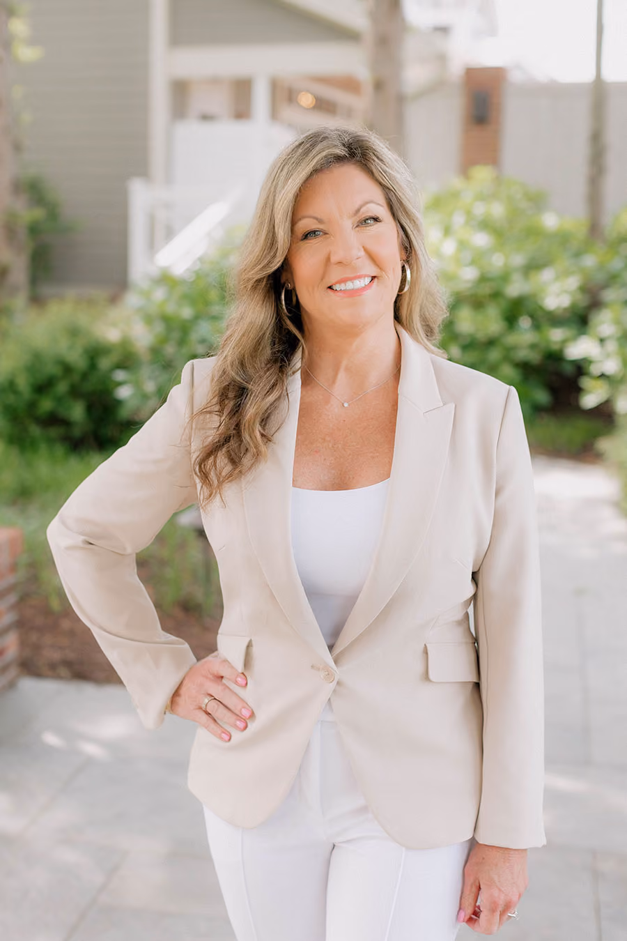 Woman with long hair in a light blazer and white top standing outdoors, smiling with one hand on her hip.