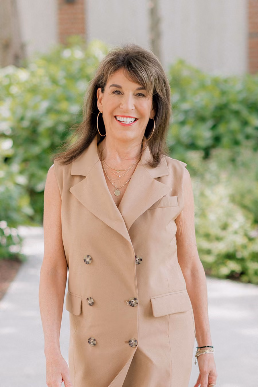 A woman in a tan sleeveless blazer dress stands outdoors, smiling, with greenery in the background.