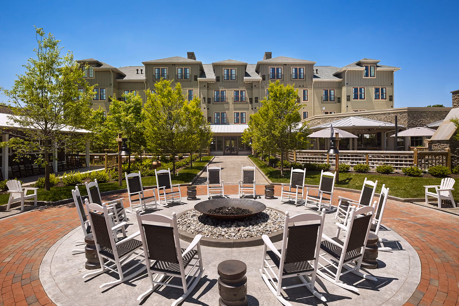 A circular arrangement of rocking chairs surrounds a fire pit in front of a large, multi-story building and garden.
