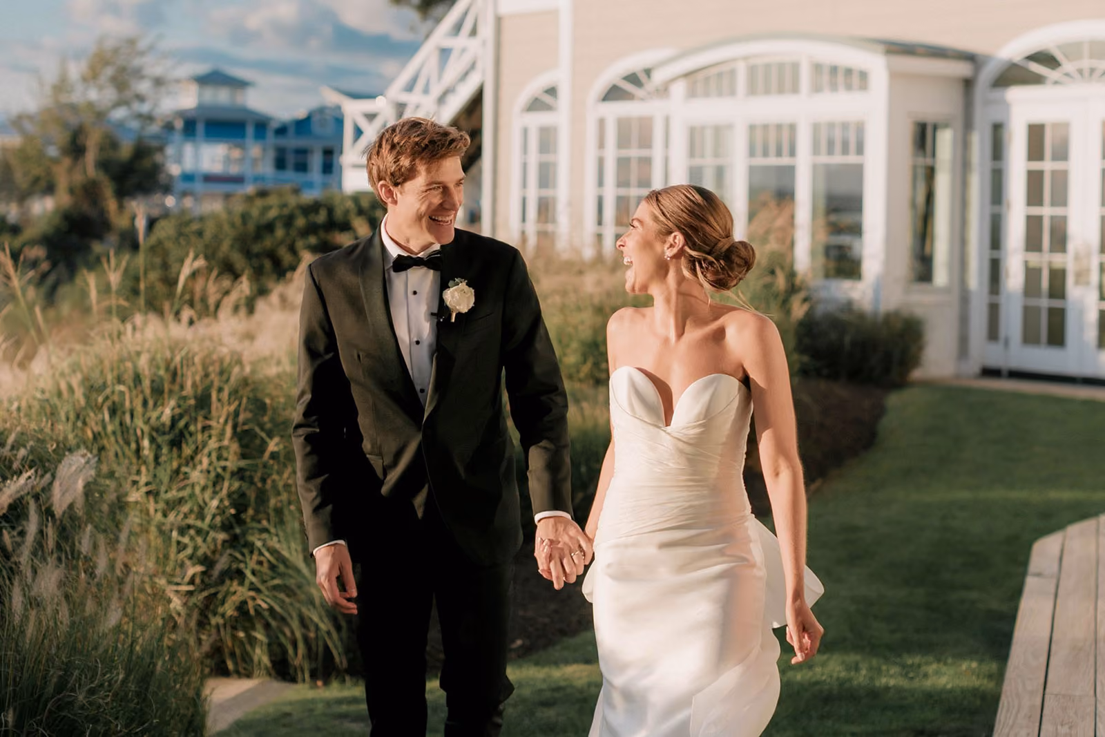 A bride and groom in formal attire hold hands and smile at each other while walking outdoors near a building.