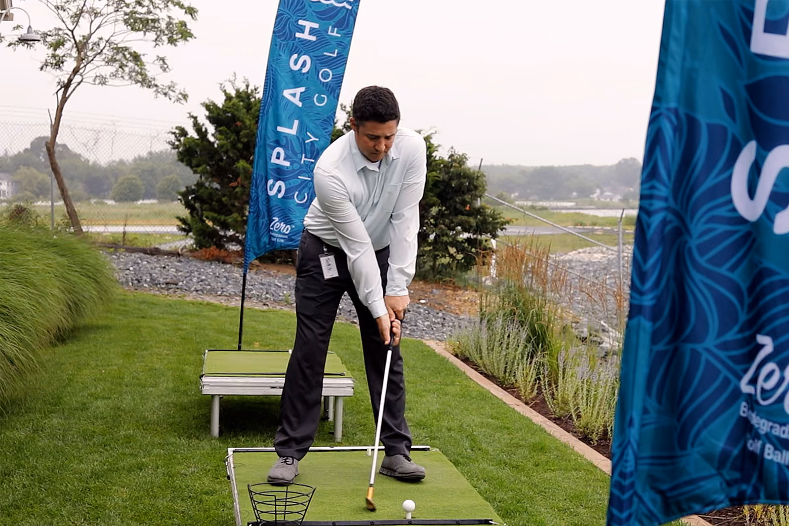 A man in business attire prepares to hit a golf ball on a small putting green with promotional flags nearby.