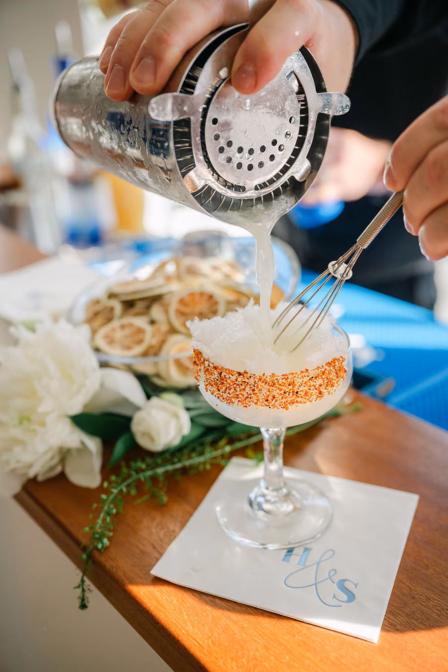 A bartender pours a cocktail into a glass with a chili-rimmed edge beside a plate of dried citrus slices.