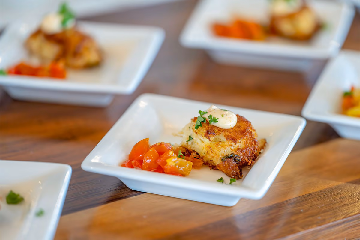 A small plate with a crab cake topped with sauce and herbs, served with diced tomatoes on the side.