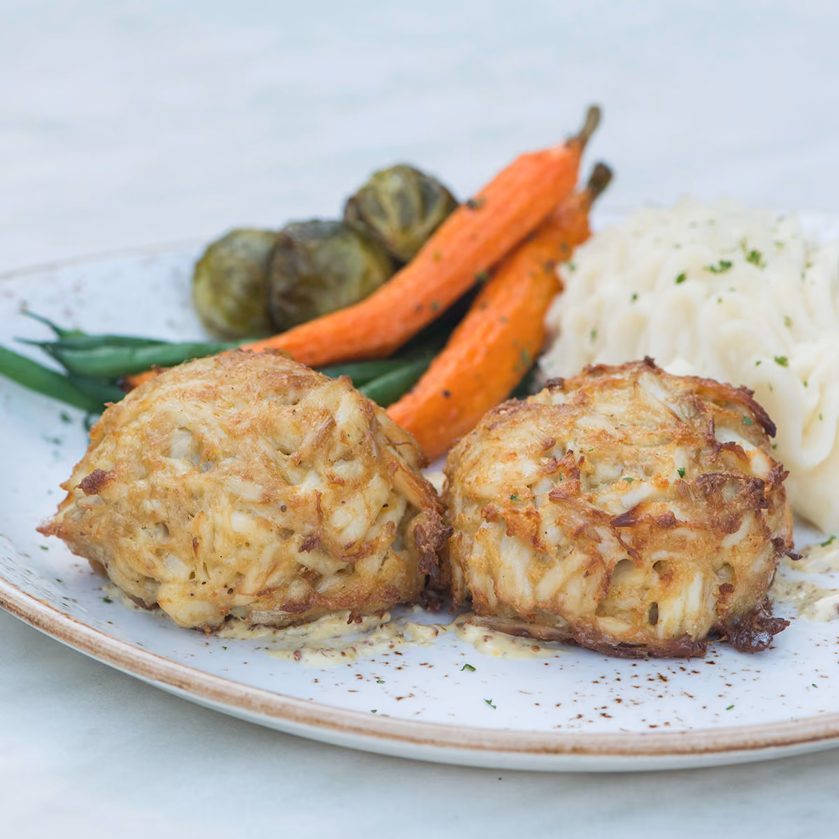 Plate with two golden-brown crab cakes served with mashed potatoes, roasted carrots, green beans, and Brussels sprouts.