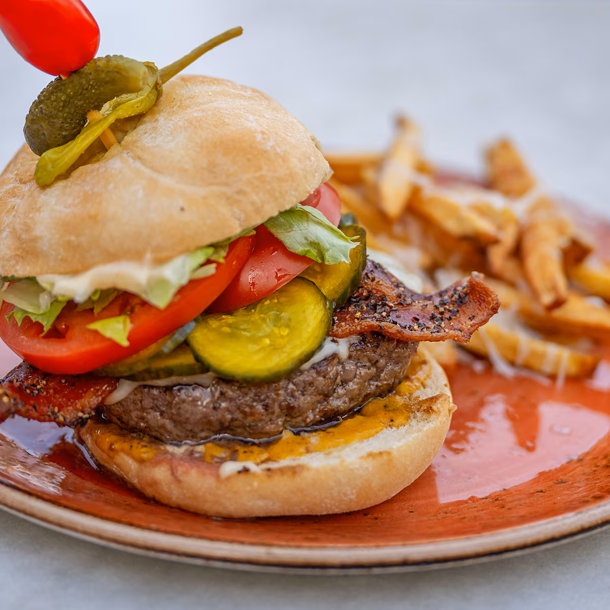 Bacon cheeseburger with lettuce, tomato, pickles, and a dollop of sauce on a bun, served with a side of French fries on a red plate.